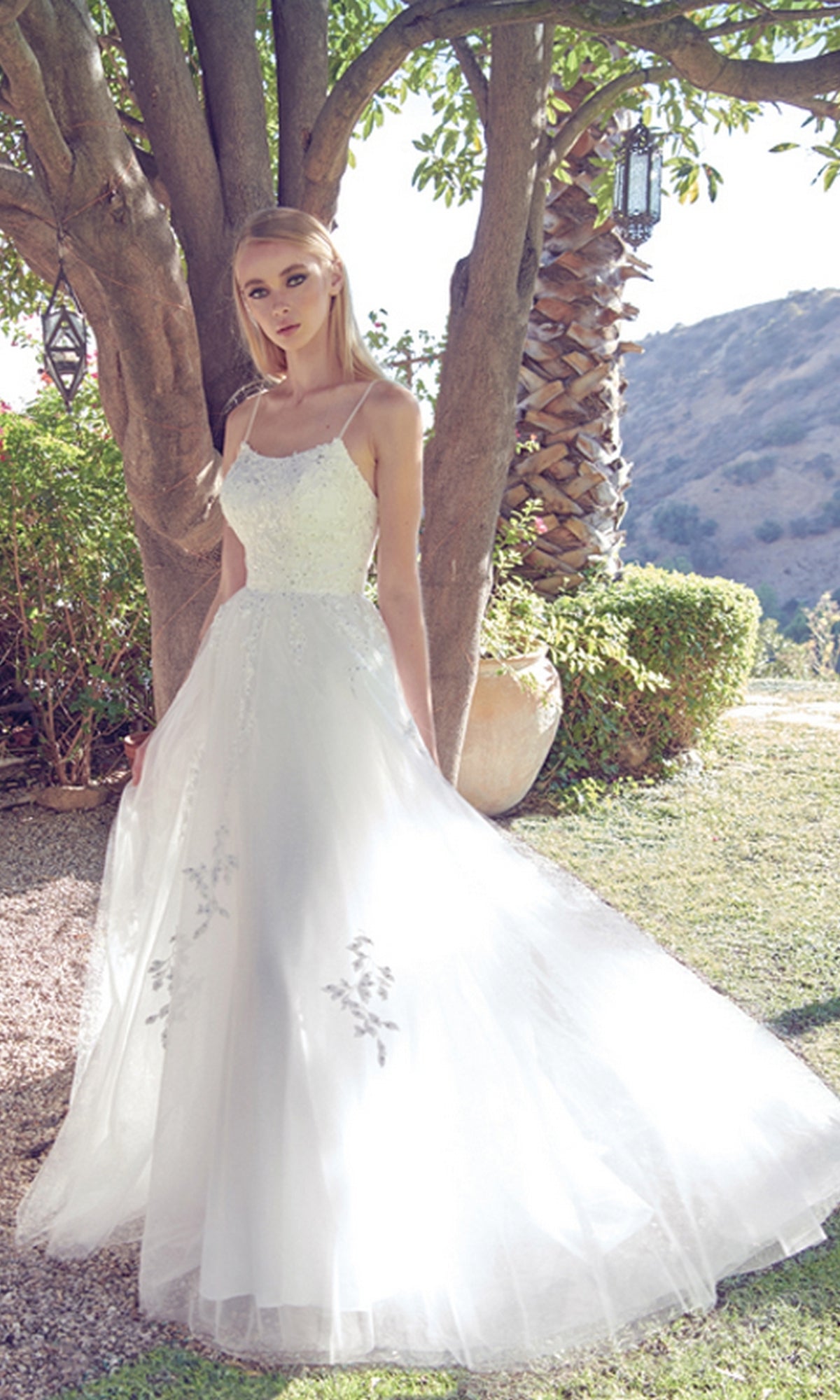 A woman wearing the Corset-Back Long Glitter-Tulle Prom Ball Gown 260, a white sleeveless formal dress with floral details, stands outdoors near a tree and greenery.